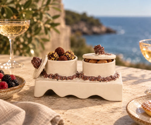 A ceramic appetizer tray with two small cream-colored bowls filled with chocolate truffles and cookies, set on a stone table with a blurred coastal background and wine glasses nearby.