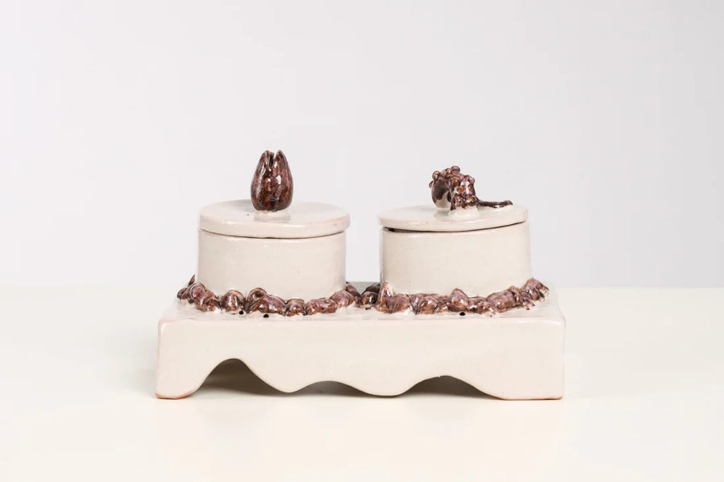 A ceramic double-candle holder set on a white background, featuring two cream-colored cylindrical candle jars with lids, decorated with dark brown textured details on the base and lid tops.
