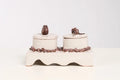 A ceramic double-candle holder set on a white background, featuring two cream-colored cylindrical candle jars with lids, decorated with dark brown textured details on the base and lid tops.