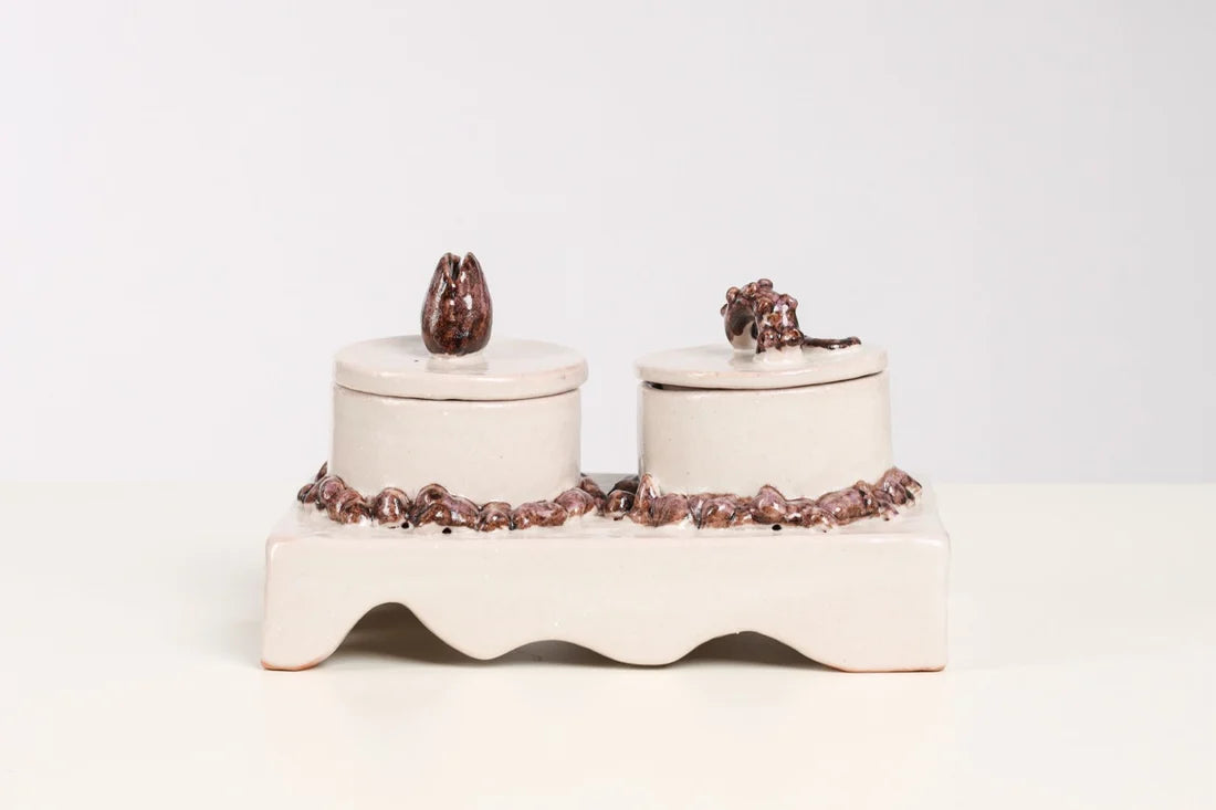 A ceramic double-candle holder set on a white background, featuring two cream-colored cylindrical candle jars with lids, decorated with dark brown textured details on the base and lid tops.