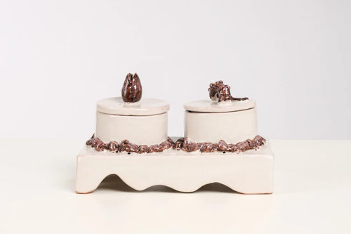 A ceramic double-candle holder set on a white background, featuring two cream-colored cylindrical candle jars with lids, decorated with dark brown textured details on the base and lid tops.