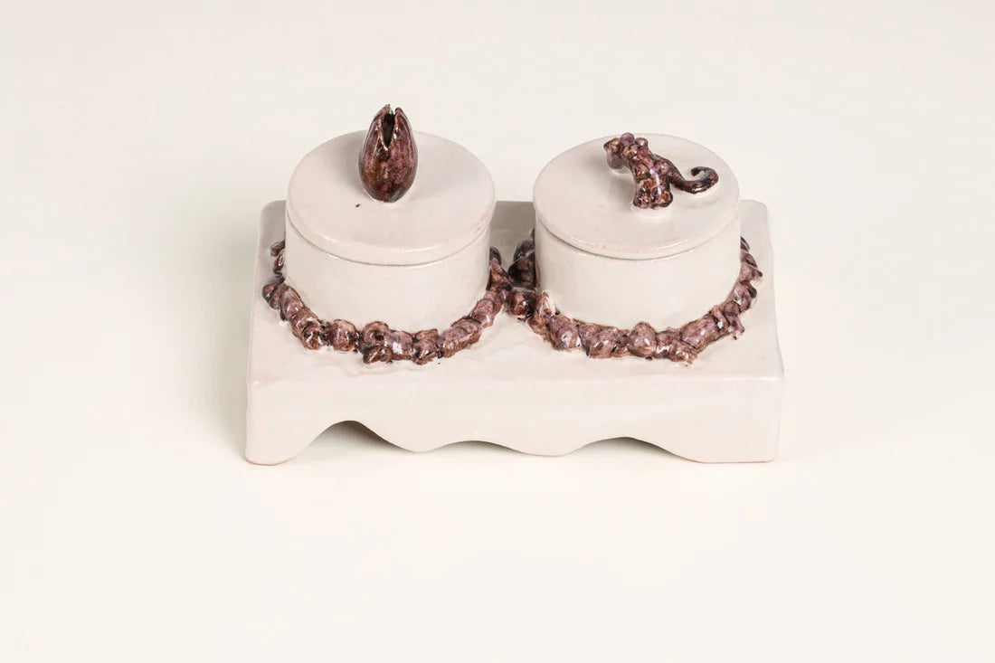 A ceramic double-lidded box set on a matching base, featuring a glossy off-white glaze and dark metallic rose-gold or bronze beaded trim with floral-shaped lids.