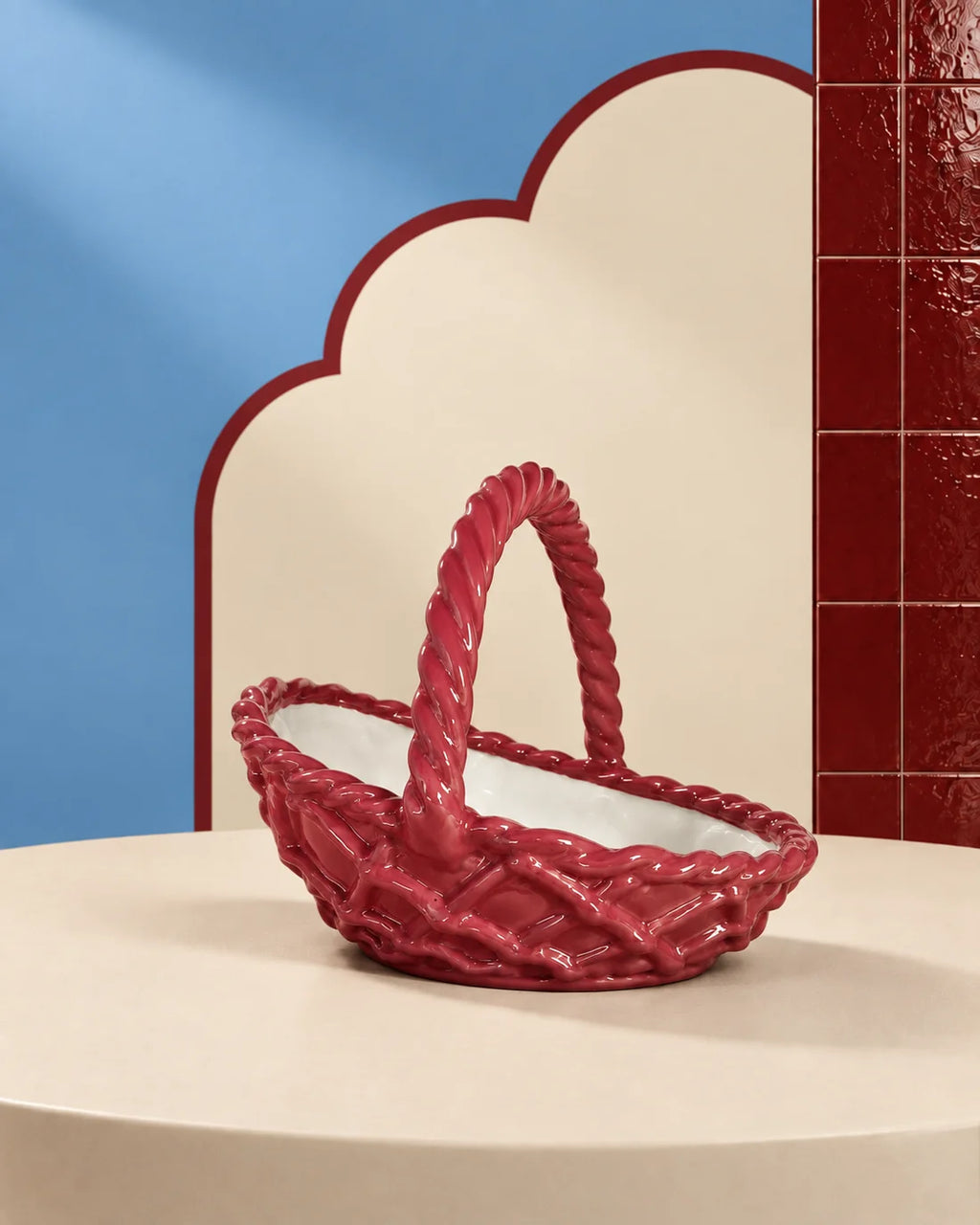 A glossy red woven basket with a twisted rope handle, displayed on a cream-colored pedestal against a stylized studio background with blue and red panels.
