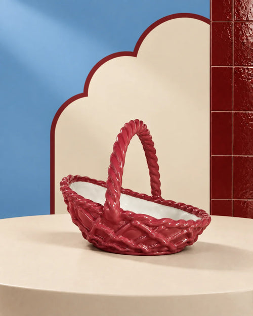 A glossy red woven basket with a twisted rope handle, displayed on a cream-colored pedestal against a stylized studio background with blue and red panels.