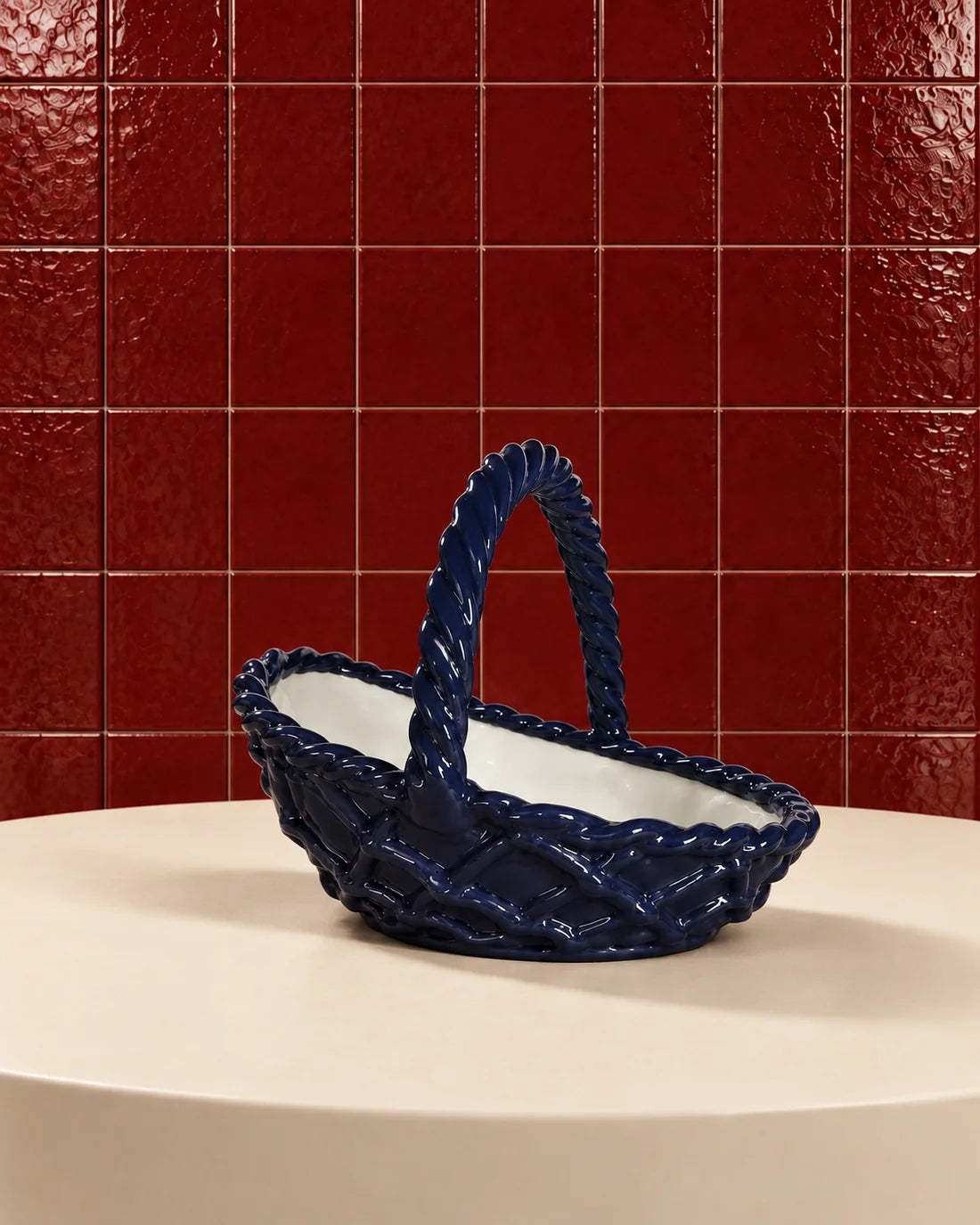 A glossy dark blue ceramic basket with a twisted handle and white interior, displayed on a cream-colored pedestal against a red tiled background.