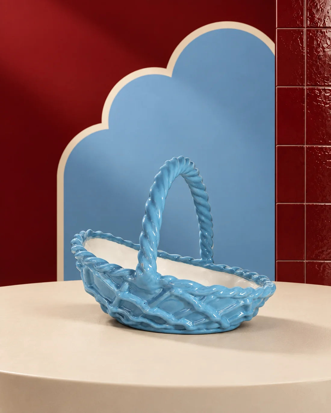 A glossy light blue ceramic basket with a twisted rope handle, displayed on a cream-colored pedestal against a stylized background of red and blue panels.