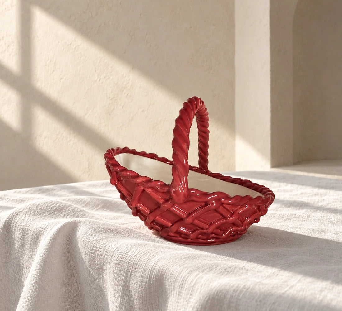 A glossy red decorative basket with a twisted handle, styled on a draped white linen cloth in warm, sunlit sunlight against a textured beige wall.