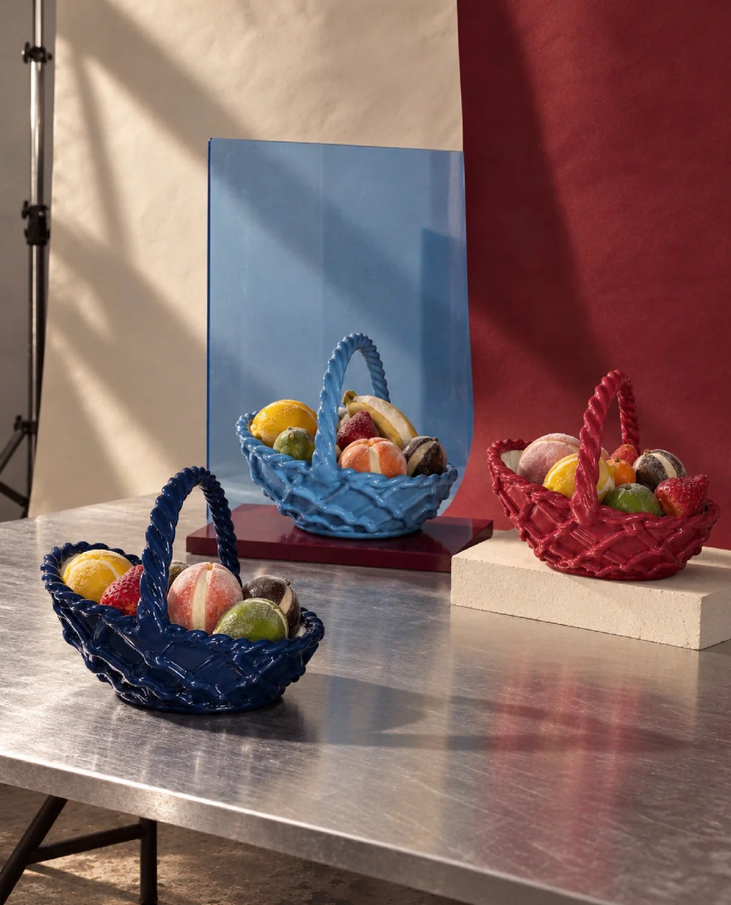 Three decorative woven baskets in blue, red, and dark blue, each filled with colorful fruits, arranged on a metallic table with a blue and red backdrop.