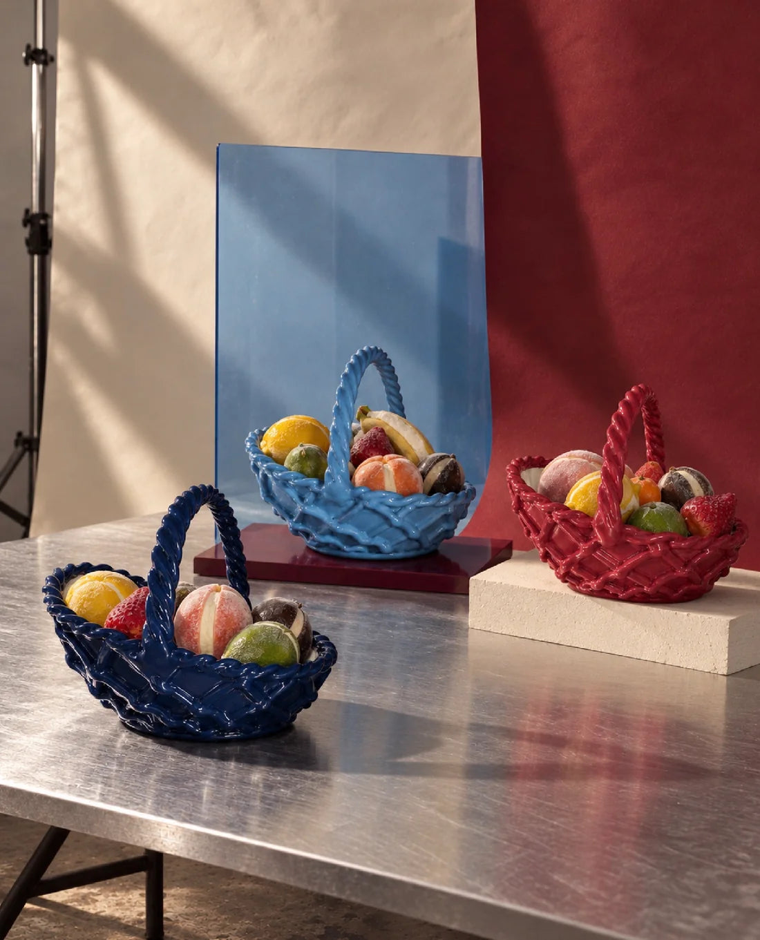 Three decorative woven baskets in blue, red, and dark blue, each filled with colorful fruits, arranged on a metallic table with a blue and red backdrop.
