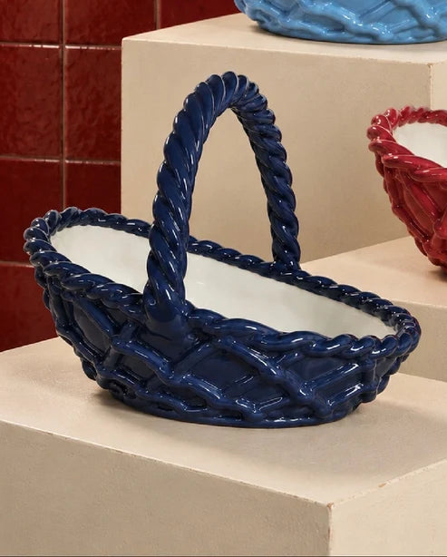 A glossy dark blue ceramic basket with a twisted rope handle and woven pattern, displayed on a beige pedestal.