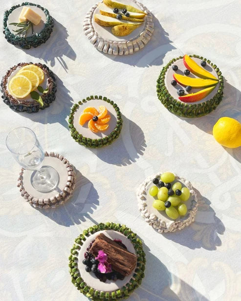 An overhead styled flat-lay of several small fruit and nut tartlets or mini cakes on a white patterned tablecloth, each bordered with green or beige pastry and garnished with citrus slices, blueberries, green grapes, or chocolate pieces. A clear wine glass sits on one tartlet.