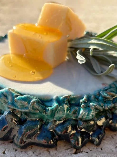 A close-up of a handmade ceramic dish featuring a textured, organic-shaped base in teal and dark blue glazes, styled with two cubes of pale yellow cheese and a drizzle of honey, with green leaves on top.