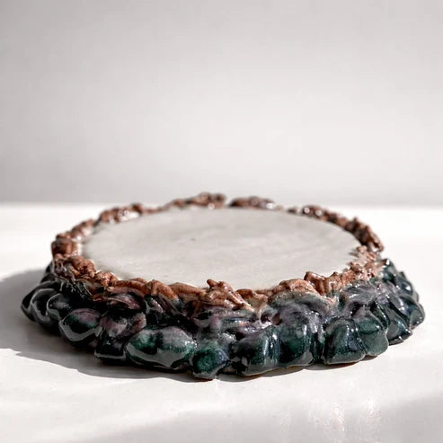 A handmade ceramic tray with a textured, organic organic-shaped base in dark green and purple glazes, and a raised brown rim, photographed on a white surface with a neutral gray background.