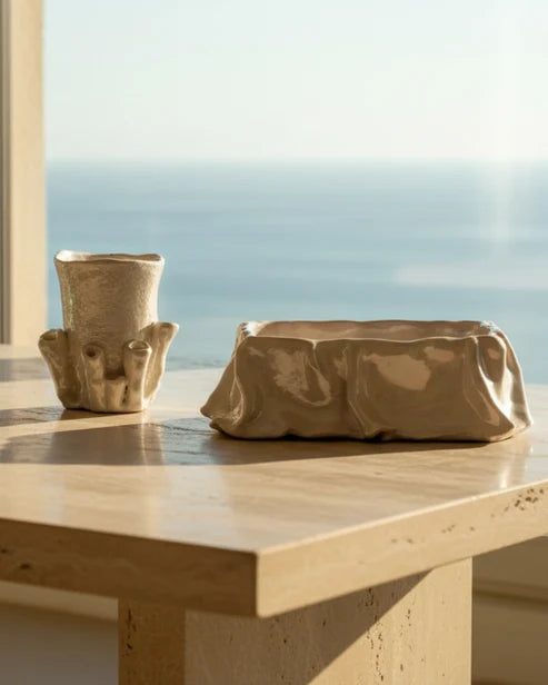 Two sculptural beige ceramic vessels, one small cup with animal-like ears and one wide shallow planter, displayed on a light stone table with a blurred ocean view in the background.