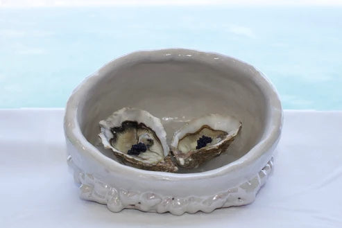 A white ceramic shell-shaped dish containing two open oysters, set on a white surface with a light blue background.