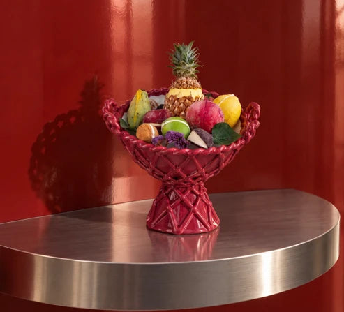 A red woven pedestal fruit bowl filled with assorted fruits, including a pineapple, displayed on a metallic silver table against a red background.