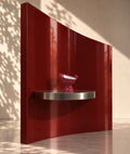 A glossy deep red curved wall-mounted shelf with a silver metal base, holding a small pink patterned bowl, set against a beige wall with dappled sunlight and shadows.