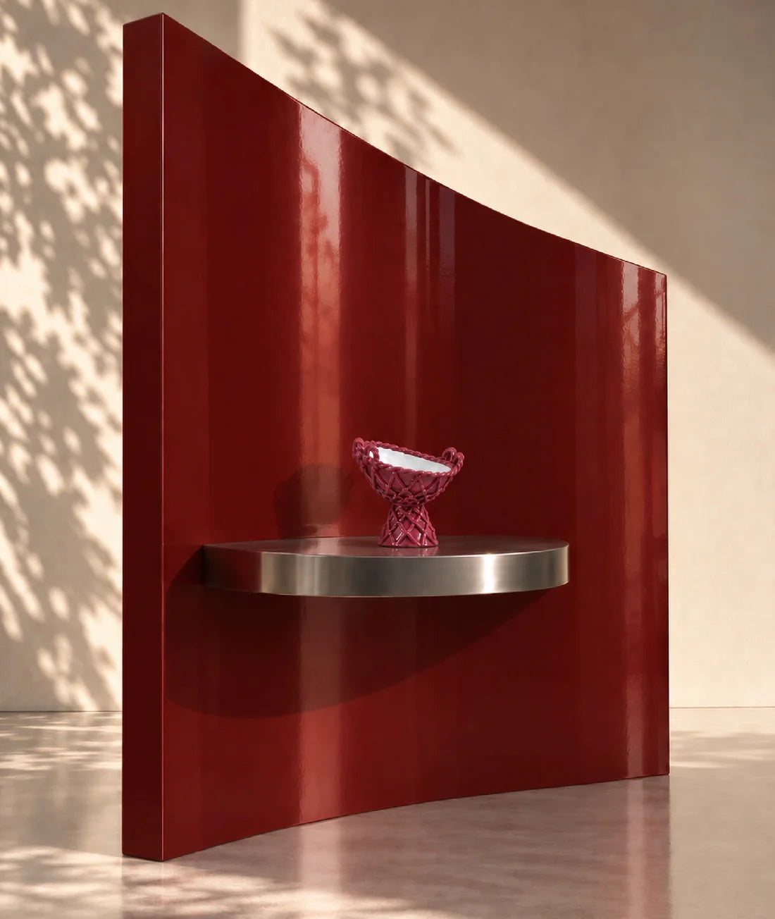 A glossy deep red curved wall-mounted shelf with a silver metal base, holding a small pink patterned bowl, set against a beige wall with dappled sunlight and shadows.