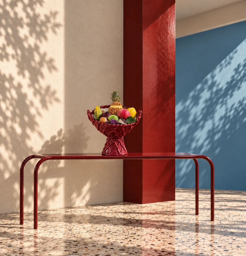 A red metal entryway table with a woven red fruit bowl on top, set in a modern architectural corner with beige and blue walls, tree shadows, and a terrazzo floor.