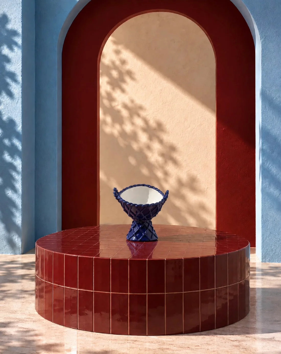A dark blue textured ceramic pedestal bowl with a white interior, displayed on a matching glossy red tiled round table in front of a red and blue arched architectural backdrop.
