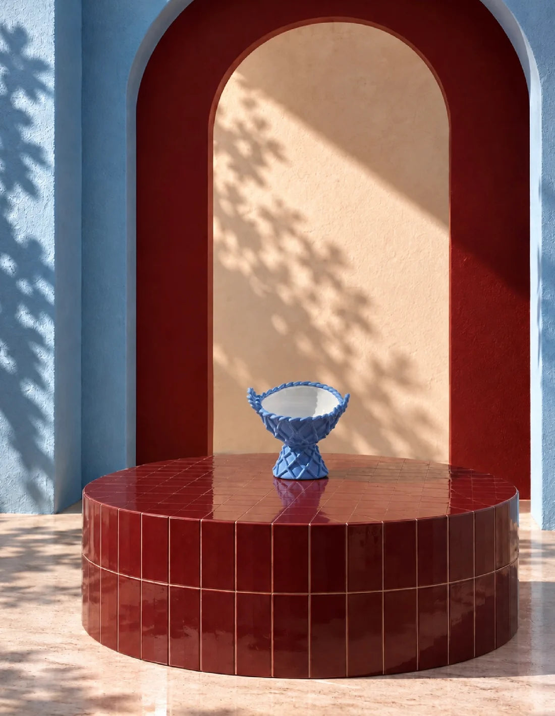A blue textured ceramic pedestal bowl sits on a glossy round red tiled table in front of a red arched alcove with blue walls and warm sunlight casting leaf shadows.