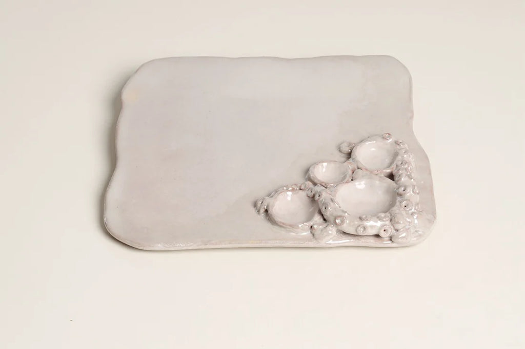 A light gray ceramic dish with a rounded rectangular shape, featuring a textured, organic cluster of small raised bowls on one side, set against a plain off-white background.