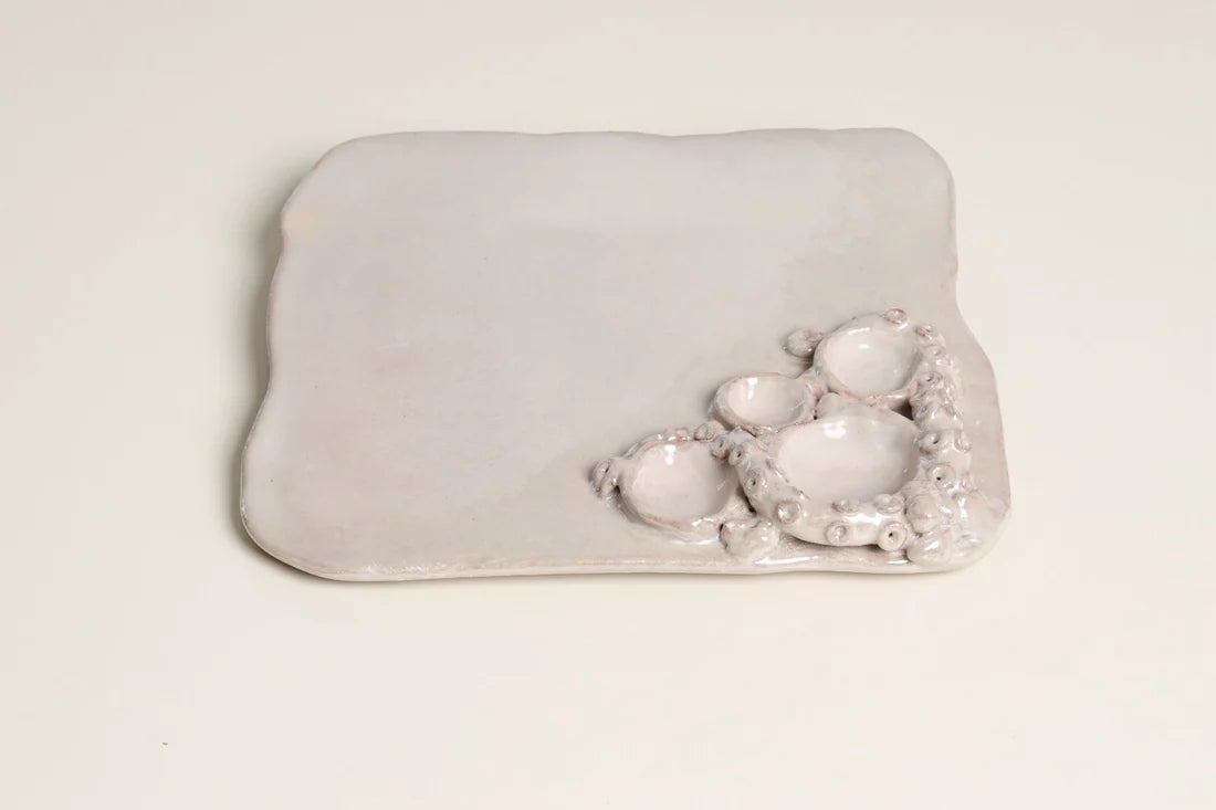 A light gray ceramic dish with a rounded rectangular shape, featuring a textured, organic cluster of small raised bowls on one side, set against a plain off-white background.