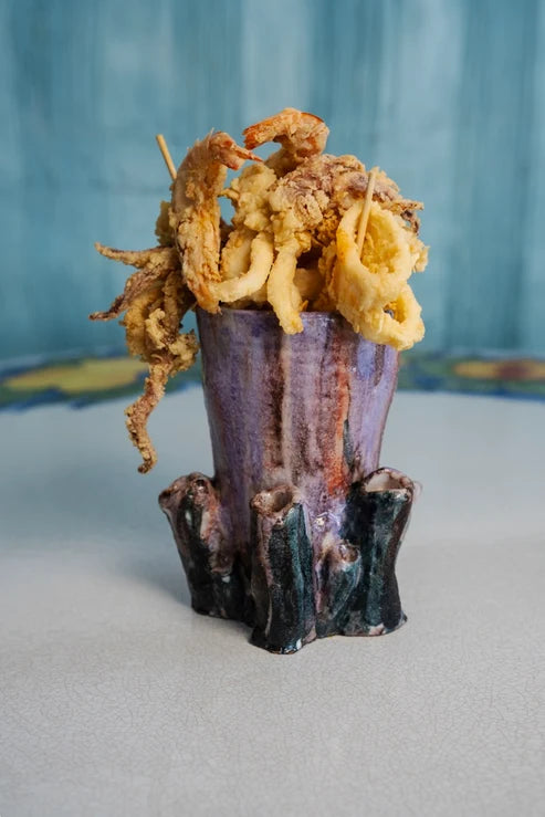 A ceramic food holder shaped like a tree trunk, filled with golden-brown fried calamari rings on skewers, set against a textured blue background.
