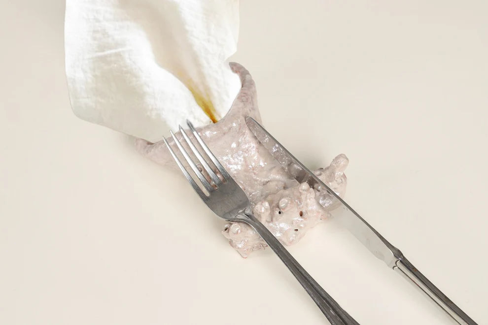 A ceramic figurine of a hand holding a knife and fork, with a white cloth draped over the top, set against a plain cream background.