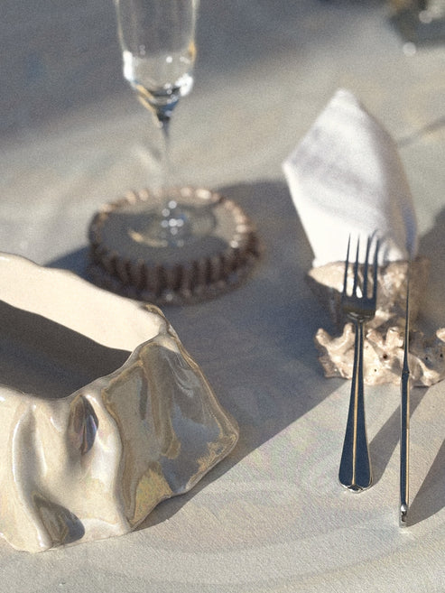 A styled tabletop scene with a white ceramic dish, a fork, a knife, a folded white napkin, and a clear drinking glass on a light surface under strong directional sunlight and shadows.