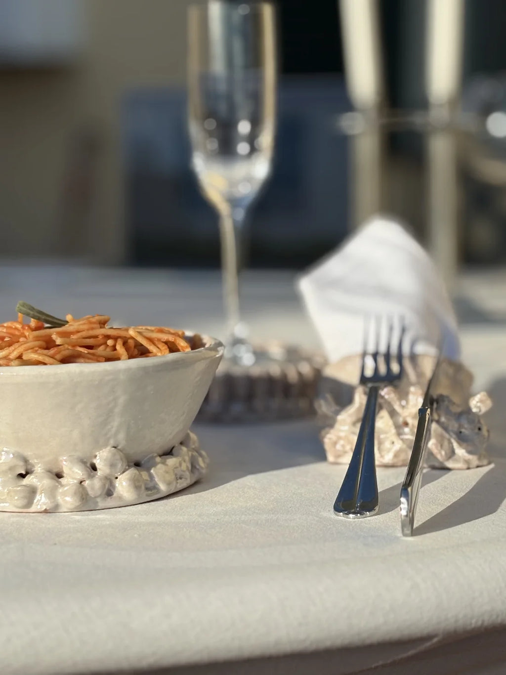A styled dining table setting featuring a white textured ceramic bowl filled with spaghetti and a small green garnish, set on a white tablecloth next to silver flatware and a folded white napkin.
