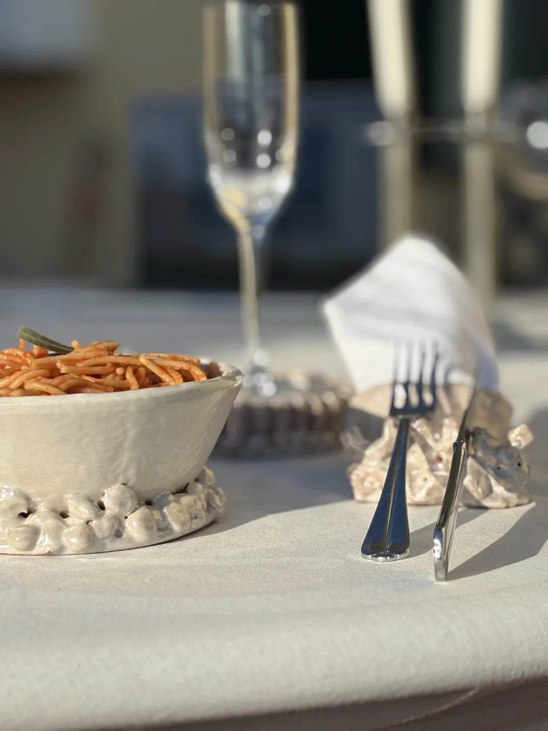 A styled dining table setting featuring a white textured ceramic bowl filled with spaghetti and a small green garnish, set on a white tablecloth next to silver flatware and a folded white napkin.